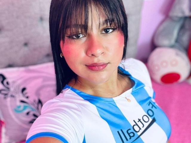SweetSoffiee with medium tits, wearing a blue and white soccer shirt, has long black hair, bangs, and a cute face, sitting on a pink bed, looking at the camera.