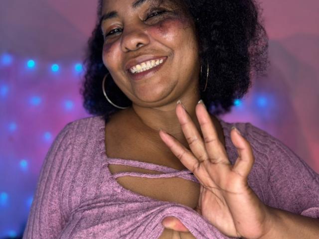 EleonorEbony, souriant avec ses cheveux noirs et bouclés, portant un haut tricot rose-violet, laissant entrevoir sa main et ses petites seins grâce à des découpes, avec des boucles d'oreilles en forme de cerceaux, un blush rose sur les joues, dans une lumière colorée..