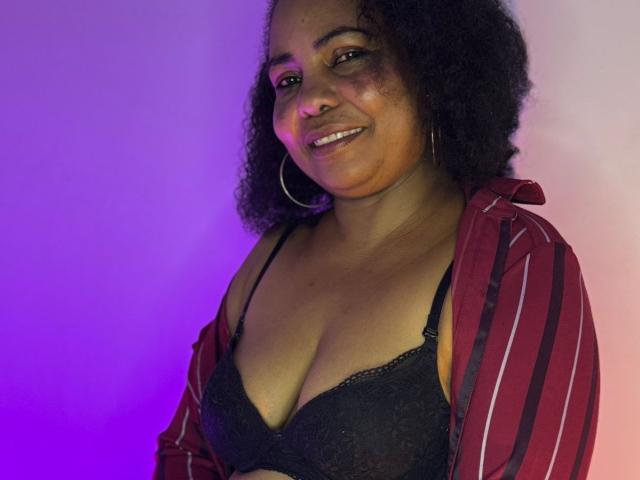 EleonorEbony, con i capelli neri ricci, indossa una camicia a righe rosse sopra un reggiseno nero in pizzo, sorridendo, con orecchini a cerchio, illuminazione viola e rosa, che mostra il seno e un viso carino..