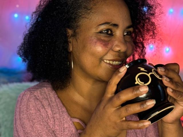 EleonorEbony, con i capelli neri ricci, la pelle marrone e un sorriso, tiene in mano una tazza di ceramica nera con decorazioni dorate, indossa un maglione rosa e orecchini a cerchio argentati..