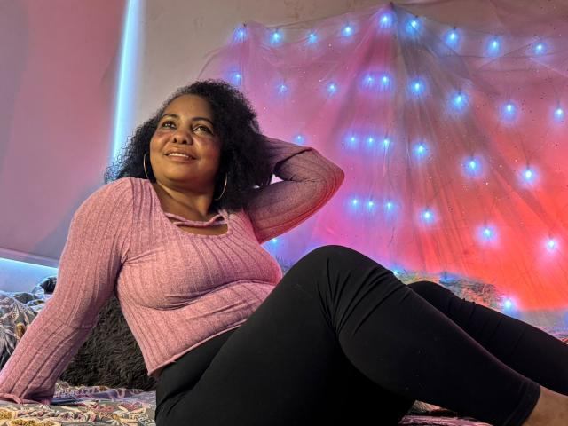EleonorEbony, donna nera formosa con seno di media grandezza, indossa una maglietta a coste rosa e pantaloni neri, seduta sul letto, che guarda verso l'alto, con capelli ricci, una mano tra i capelli, luci fatate blu e rosse..