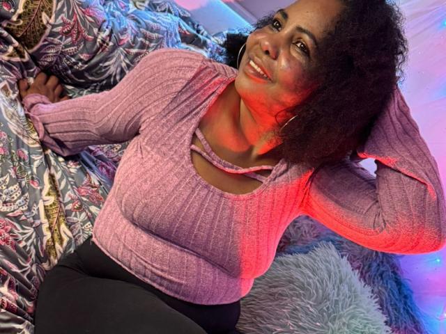 EleonorEbony, avec des cheveux bouclés, souriante, portant un haut rose côtelé et un pantalon noir, allongée sur un lit avec des draps colorés, une main dans ses cheveux, avec des lumières rouges et bleues..