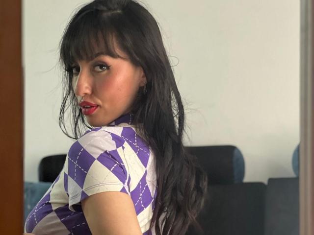 EmilyKateK with long black hair, purple and white diamond-patterned top, red lipstick, looking over shoulder, showing her tits, standing in front of a mirror, indoors.