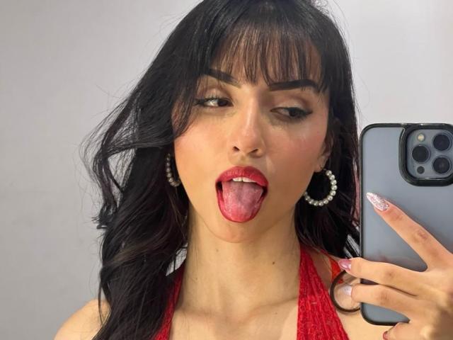 EmilyKateK with dark hair, red lipstick, and tongue out, holding a phone, wearing hoop earrings, and a red halter top, showing off her pink-tipped nails.