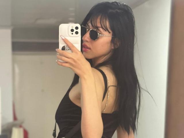 EmilyKateK with long black hair, wearing sunglasses and a black dress, taking a mirror selfie with a white iPhone. Her tits are visible through the dress's thin straps.