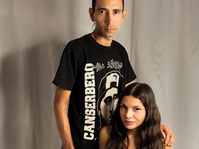 EdieAndLily with short dark hair, wearing a black t-shirt, stands behind Lily with long dark hair, smiling, both touching shoulders, in a studio setting.