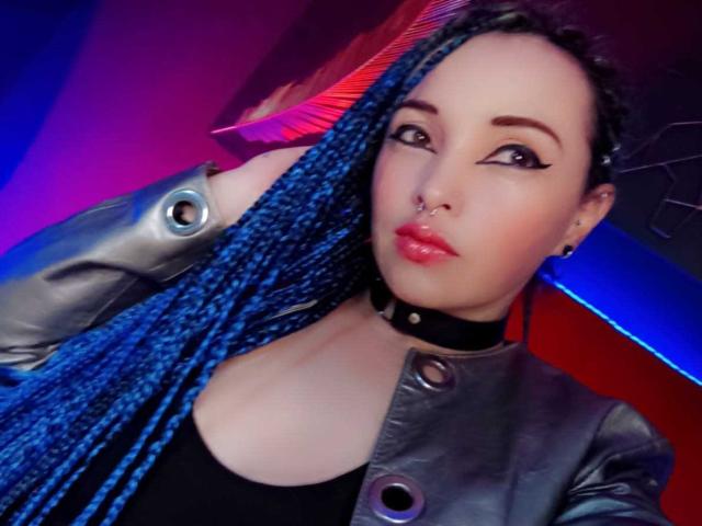 DalilaMusa with blue braids, wearing a black choker, silver jacket, and black top, showing off her tits, nose ring, and glossy pink lips, with a confident, sexy look.