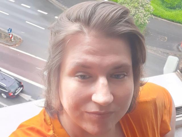 Beastbitch-hot has a slightly puffy nose, light brown eyes, and short brown hair, wearing an orange shirt, with a car and road in the background.