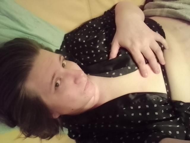 Beastbitch-hot lying on a couch, wearing a black, patterned shirt, with her hand on her chest, exposing her bare, small tits, and a slight tan line.