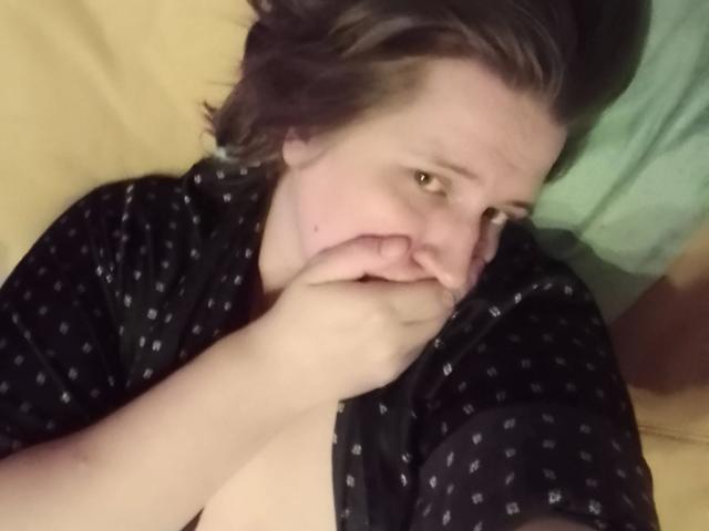 Beastbitch-hot lying on a bed, covering her mouth with her hand, wearing a black polka dot shirt, brown hair, looking up with a slightly tired expression.