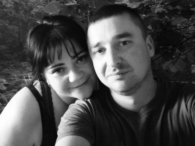 DebbieXRoy with short dark hair and bangs, smiling, leans on a guy with a shaved head and light beard, wearing a dark shirt. Black and white selfie.