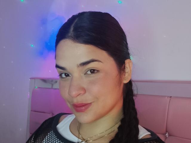 AntonelaPark has dark hair in a braid, brown eyes, light skin, and red lipstick. She's wearing a black mesh top with a white shirt underneath. Her ass is not visible.