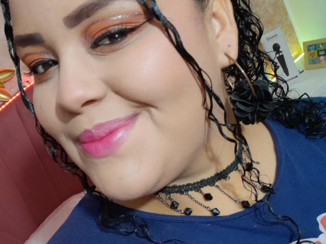 Melannie Carter, con labbra rosa, trucco marrone per gli occhi, trecce nere, camicia blu, collana nera e orecchini, sorride leggermente, mostrando il suo sedere prosperoso nella parte inferiore dell'immagine..