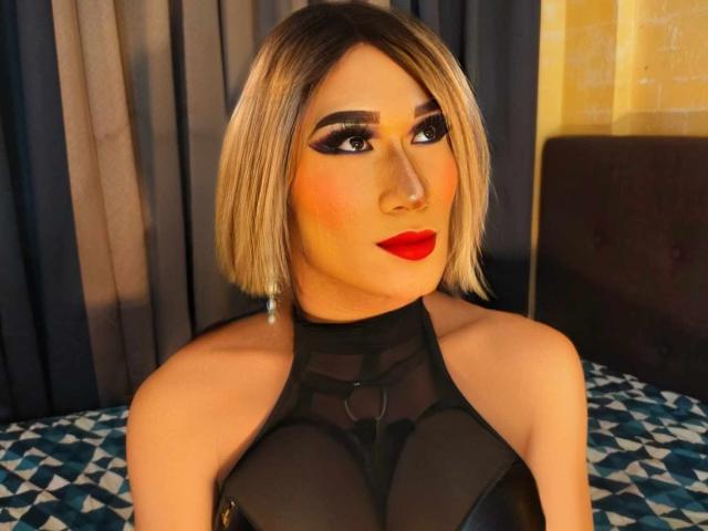 Hotjessicatrans con un corte de pelo rubio, delineador de ojos negro, lápiz labial rojo y una blusa negra transparente que muestra los pezones. Está mirando a la cámara con una ligera sonrisa. El escenario es un dormitorio, con una cama de patrón azul..
