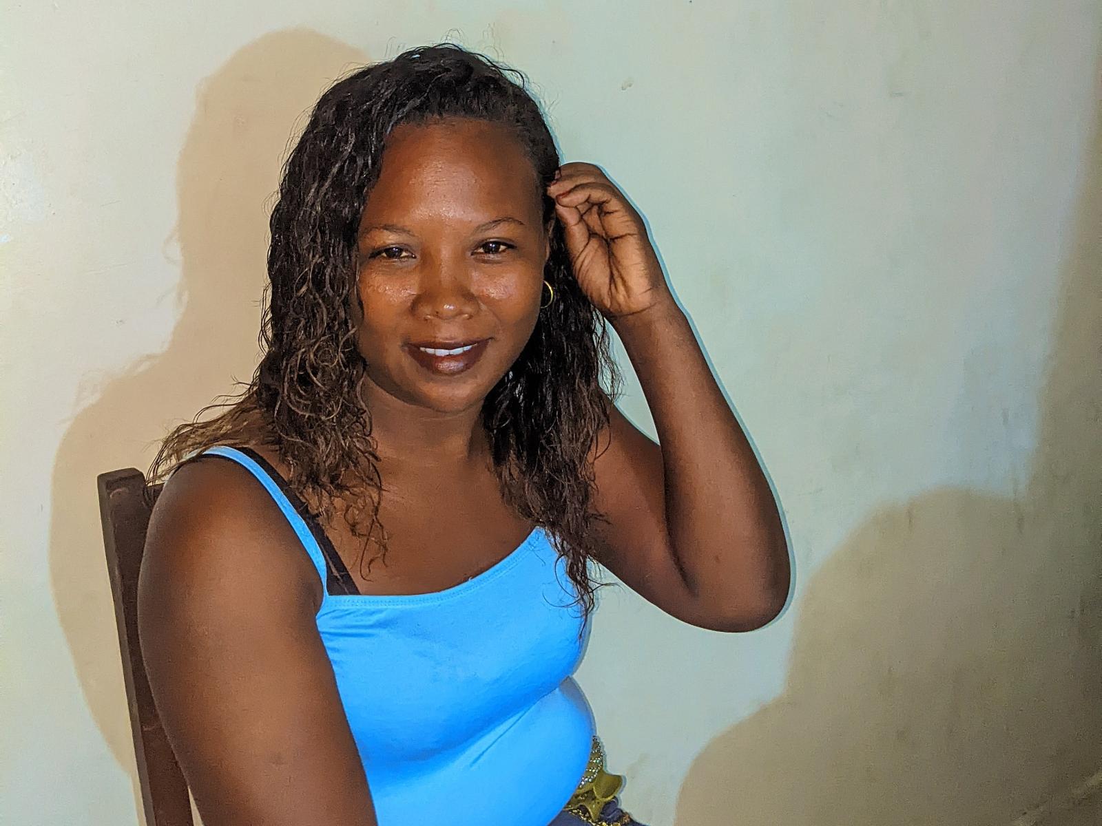PrettyMargaret, avec sa peau foncée, ses cheveux bruns bouclés, portant un débardeur bleu, touchant sa tête, souriant, montrant son bandeau de soutien-gorge noir, assise sur une chaise en bois..