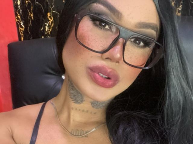 DannikaSaenz with large glasses, pink lipstick, and tattoos on her neck, looking at the camera with a slightly open mouth. She has black hair and freckles.