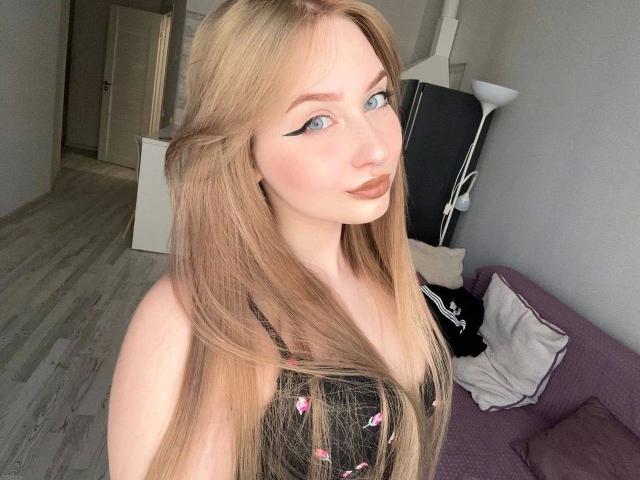 KittenKiss-hot with long blonde hair, blue eyes, and black dress with pink gems, showing off her tits, standing in a room with a purple couch and white floor lamp.