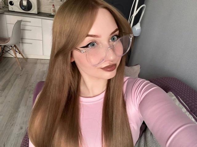 KittenKiss-hot with long brown hair, pink glasses, and pink top, showing her ass with a cute face, standing in a room with light wood flooring and white cabinets.