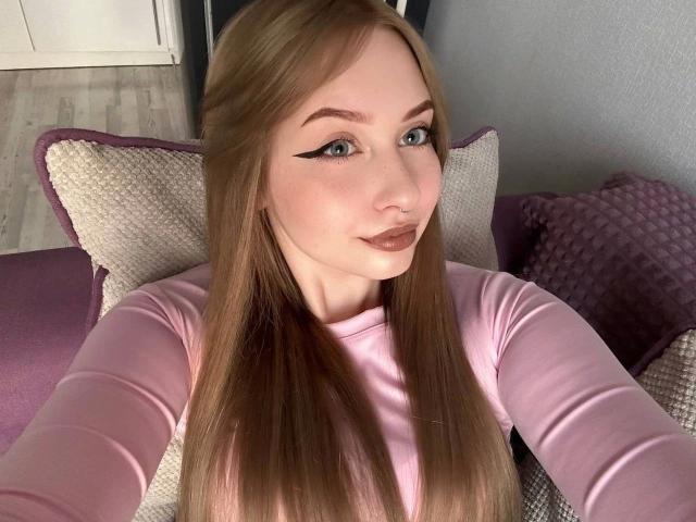 KittenKiss-hot with long brown hair, blue eyes, and pink eyeliner, wearing a pink top, taking a selfie. Boobs visible under top. Purple pillows in background.