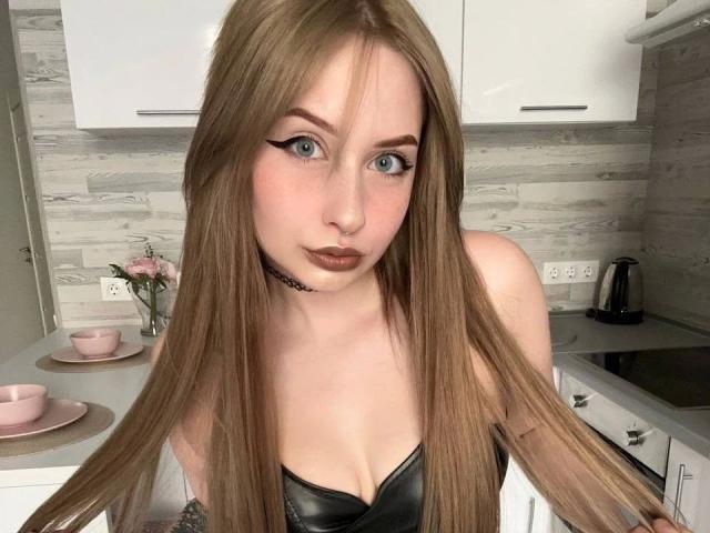 KittenKiss-hot with long brown hair, blue eyes, black leather top, choker, and brown lipstick, holding hair, in a kitchen with pink cups and white cabinets.