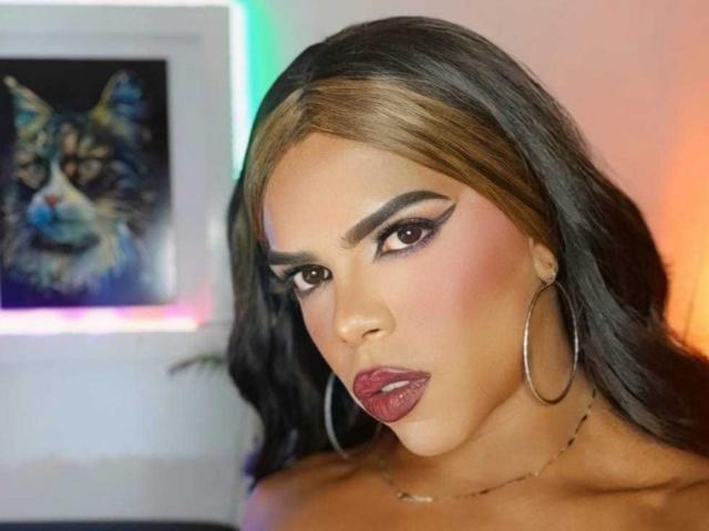 MistressCumx with long brown hair, hoop earrings, and bold makeup, has glossy red lips and a serious expression. Her dark eyes and heavy eyeliner stand out.