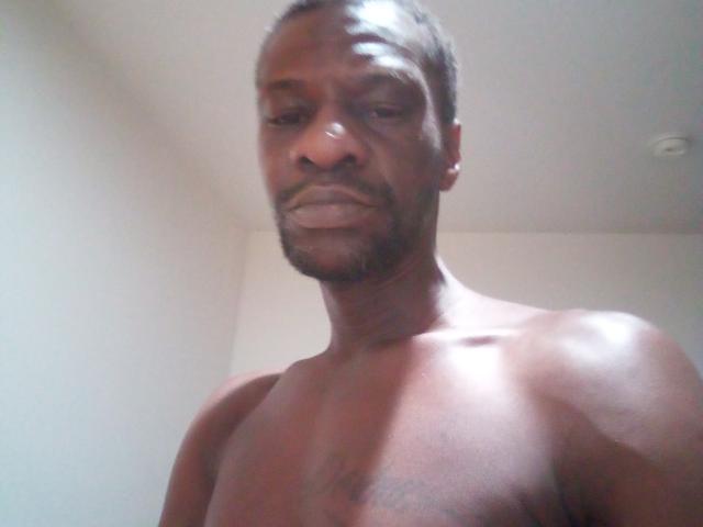 ZillaNasty is a dark-skinned, muscular man with short black hair, a beard, and a tattoo on his chest, standing shirtless and looking down at the camera.