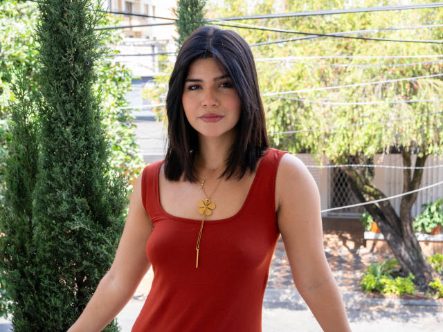 ScarlletZhen with dark brown hair, red tank top, small tits, and a gold necklace with a clover pendant, standing outside with a green tree to her left.