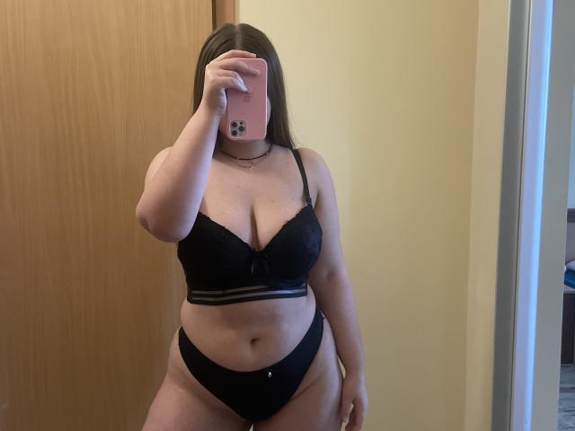 Uma mulher com seios médios, usando um sutiã de renda preta e calcinha preta, segurando um telefone rosa em frente ao rosto, em pé em um quarto com uma porta de madeira..