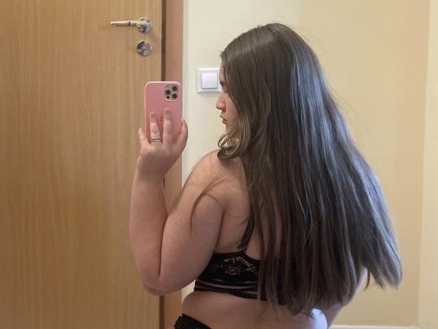 Eine Frau namens Madrinia mit langen braunen Haaren, die ein schwarzes Spitzen-BH trägt, macht ein Selfie im Spiegel. Sie hält ein rosa Telefon, zeigt ihren Rücken und ihre Brust, lächelt. Im Hintergrund sind eine Tür und ein Lichtschalter zu sehen..