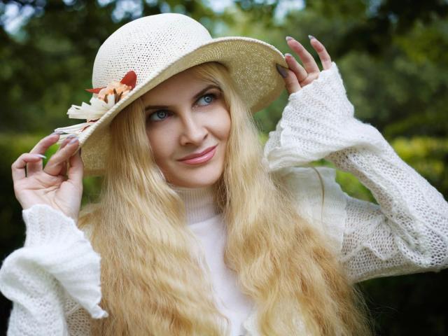 Fetish-Dreams-hot with long blonde hair, white knit sweater, and straw hat adorned with orange flowers, smiling, holding hat, blue eyes, fair skin, outdoors, soft natural light.