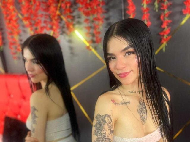 LucyLeroy mit langen, schwarzen Haaren, lächelnd, die Tattoos auf ihrer Brust und ihrem Arm zur Schau stellend, trägt ein trägerloses Oberteil, wird in einem Spiegel reflektiert, im Hintergrund sind rote Blumen zu sehen..