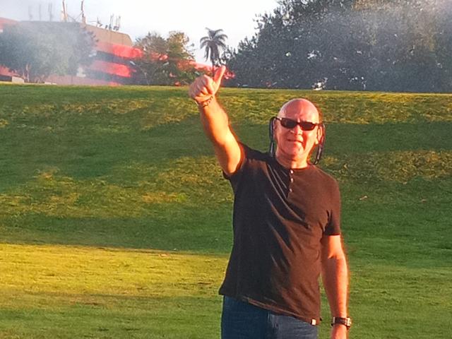 Ein Paar, der Mann mit kahlköpfigem Kopf, schwarzem T-Shirt und Sonnenbrille, zeigt mit der rechten Hand nach oben, steht auf grünem Gras, trägt blaue Jeans und eine Armbanduhr am linken Handgelenk..