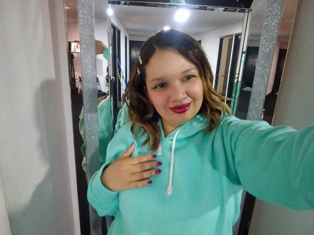 Bianca Morettii, con cabello castaño ondulado, vistiendo una sudadera verde azulada, sonriendo a la cámara, con lápiz labial rosa, uñas moradas, con una mano en el pecho, de pie frente a un espejo..