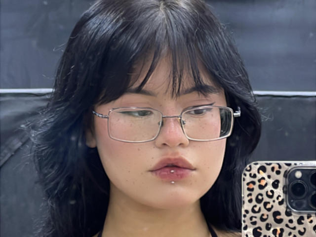 ElizabethRoux has black hair, glasses, and a pierced lower lip. She's holding a phone with a leopard print case, looking into the camera with a neutral expression.