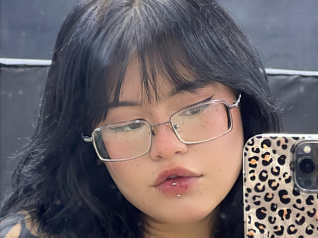 ElizabethRoux with black hair, glasses, and a lip piercing, holding a leopard-print phone case, showing her pale skin and slightly parted, pouty lips in a selfie.