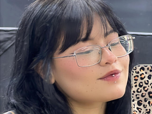 ElizabethRoux with black hair, glasses, light skin, and a lip piercing, holding a phone with a leopard print case, smiling slightly.