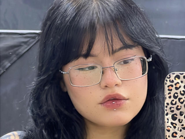 ElizabethRoux with black hair, glasses, and a lip piercing, holding a leopard-print phone case, has freckles and a slightly pouty expression, looking into the camera.