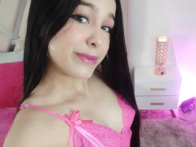 DulceTernura with long black hair, pink lipstick, and pink lace lingerie showing her tits, smiling at the camera. Pink lamp on white nightstand in the background.