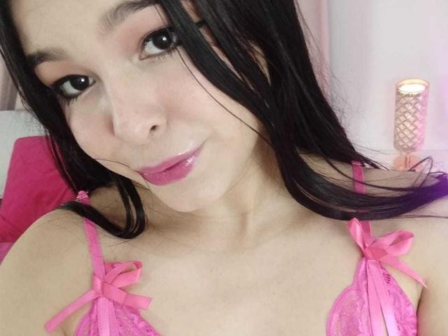 DulceTernura, com batom rosa, cabelo preto e um sutiã de renda rosa com fitas, sorrindo para a câmera. Seus seios estão visíveis no sutiã de renda..