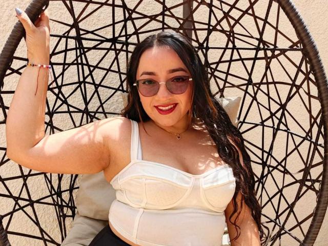 ViiolettRosse, con grandi seni, capelli neri ricci, che indossa occhiali da sole, un reggiseno bianco, un perizoma nero, sorridente, seduta su una sedia a dondolo nera in vimini, con la mano destra sollevata..