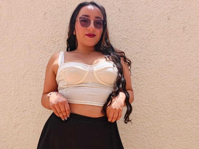 ViiolettRosse, con cabello negro y rizado, vestida con un top de encaje blanco que deja ver parte del busto y una falda negra, de pie frente a una pared beige con textura, mirando a la cámara con lápiz labial rosa..