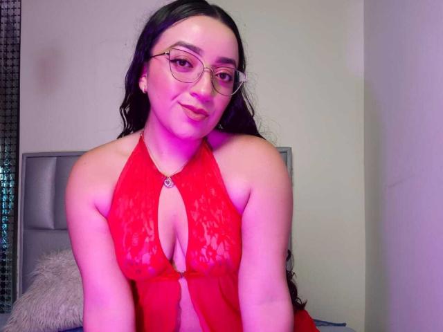 ViiolettRosse con gafas, vistiendo un top de encaje rojo con tirantes que deja ver sus pechos, sentada y sonriendo a la cámara, en una habitación con luz tenue..