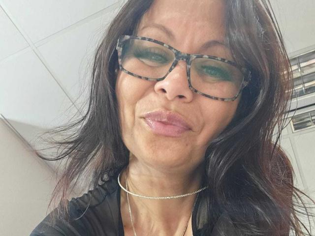 ColetteBlanche has dark brown hair, green-tinted glasses, and glossy pink lips, wearing a black top with a thin silver necklace. Her face is slightly wrinkled.