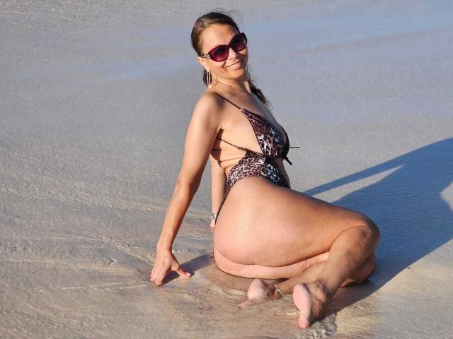 ColetteBlanche is kneeling in wet sand, wearing a leopard print bikini, showing her ass and thick thighs, with sunglasses and a smile, her back to the camera.