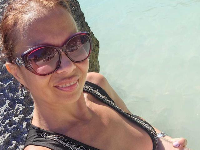 ColetteBlanche with big red sunglasses, smiling, showing her tits in a black top, standing by rocky water, brown hair in a bun, sun shining, looking cute.