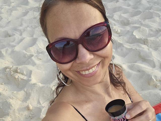ColetteBlanche with brown hair, wearing large red sunglasses, smiling, holding a small black cup, and wearing large silver hoop earrings, on a sandy beach.