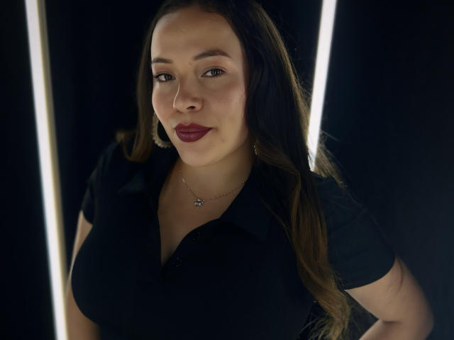 HoneyHotX with large tits, wearing a black shirt, long brown hair, red lipstick, and a small star necklace, standing in front of bright vertical lights.