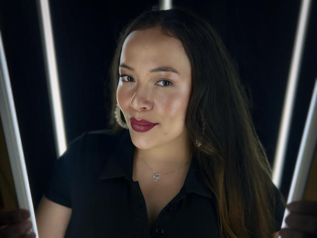 HoneyHotX mit dunkelbraunen Haaren, rotem Lippenstift und einem schwarzen Hemd, die vertikale Stäbe hält, eine Sternkette trägt und leicht lächelt. Sie hat ein süßes Gesicht und eine zarte Haut..