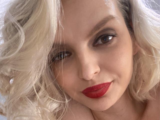 DouceAmelia's face with curly blonde hair, red lipstick, and a slight smile, brown eyes, and fair skin in a close-up shot. Her tits are partially visible at the bottom.