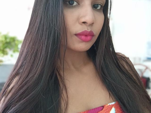 Giuliabe has long, straight brown hair, pink lipstick, and a strapless top with colorful patterns. She has tan skin and blue eyes, looking at the camera with a serious expression.